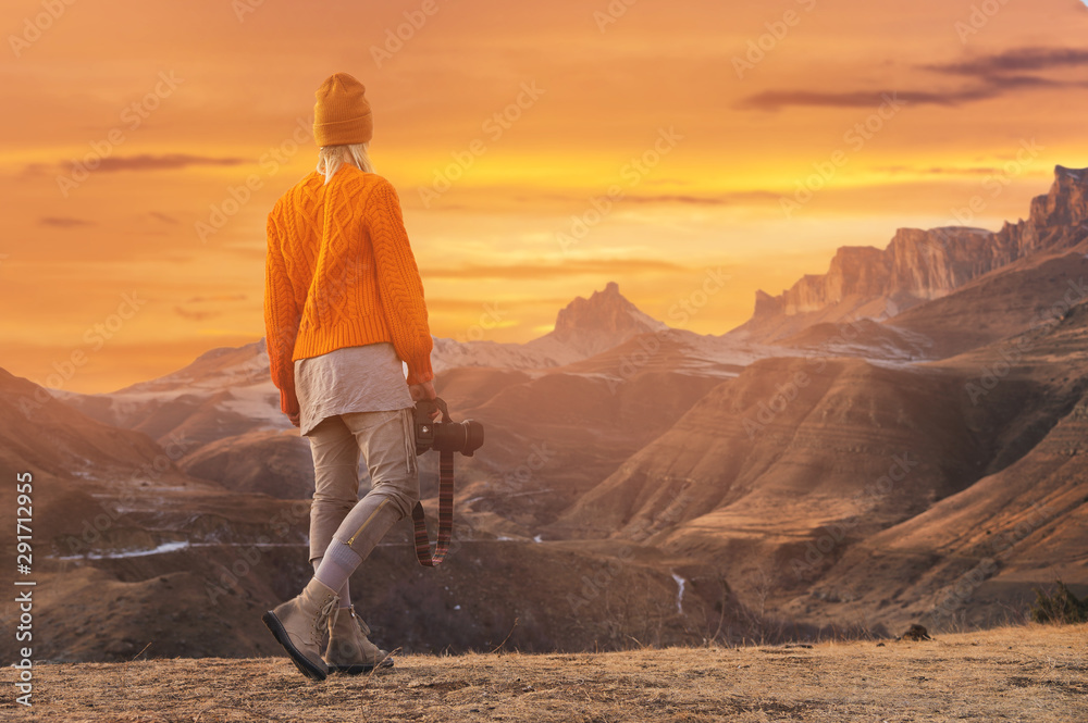 Portrait from the back of the girl traveler photographer in an orange sweater and hat with a camera in hand in the mountains against the background of a frozen mountain lake. Photo travel concept