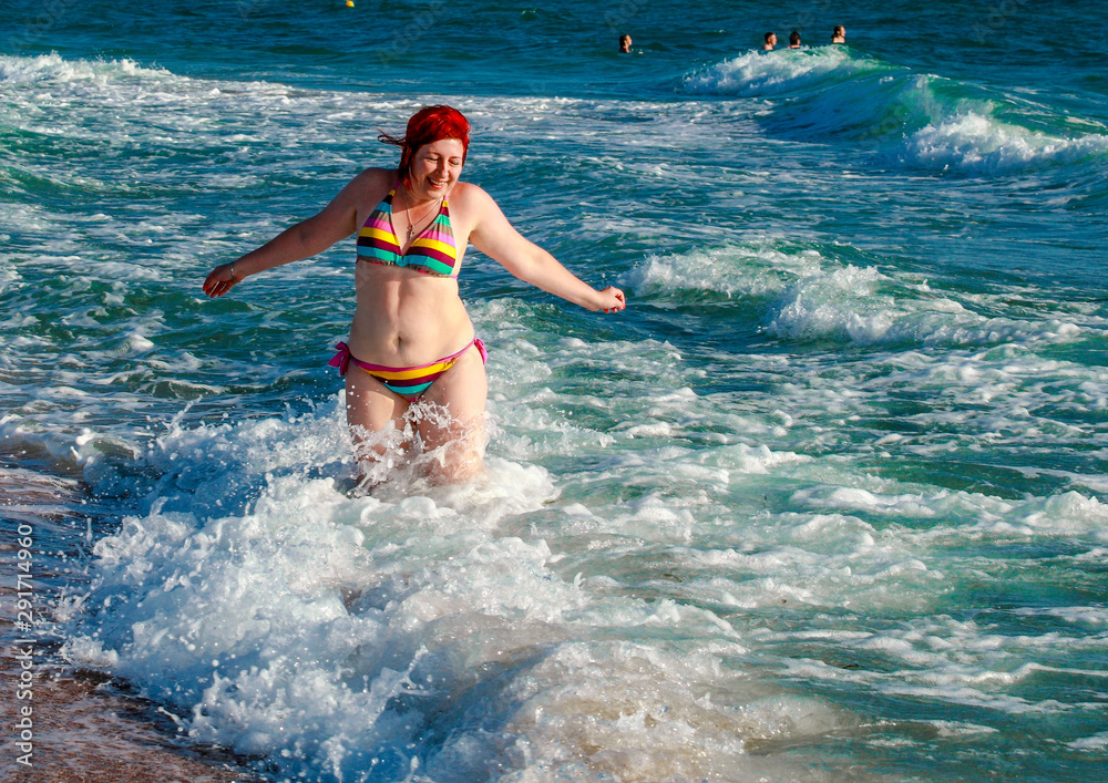 Waves break over a young woman standing in the sea. She is overflowing with happiness, laughing and enjoying life.