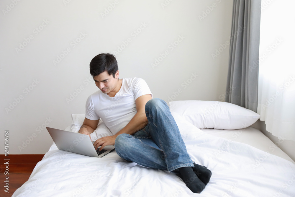 portrait of handsome caucasian in white shirt and blue jean using computer on bed in bedroom with relaxation and smiling face portrait of handsome caucasian in white shirt and blue jean using computer on bed in bedroom with relaxation and smiling face