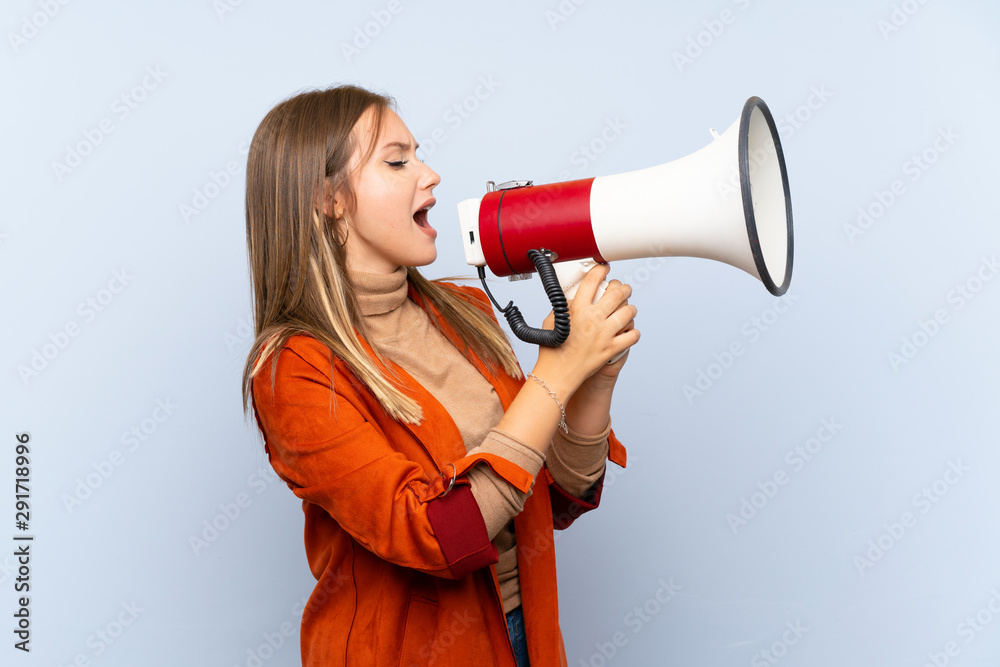 Teenager girl with coat over isolated blue background shouting through a megaphone Teenager girl with coat over isolated blue background shouting through a megaphone
