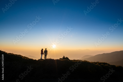 Silhoutte of a couple hold hand of each other looking at sunset on top of hill with background of mountain and golden color of sunshine. Concept of teamwork, successful.