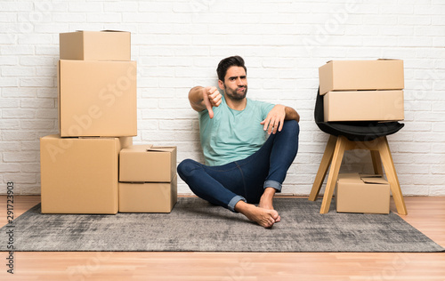 Handsome young man moving in new home among boxes showing thumb down sign