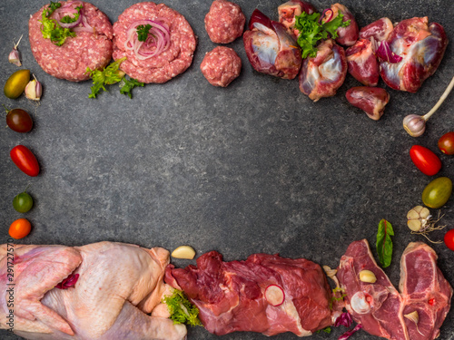 Assorted raw meat on dark background, with cherry tomatoes, garlic, onion and greens, copy space