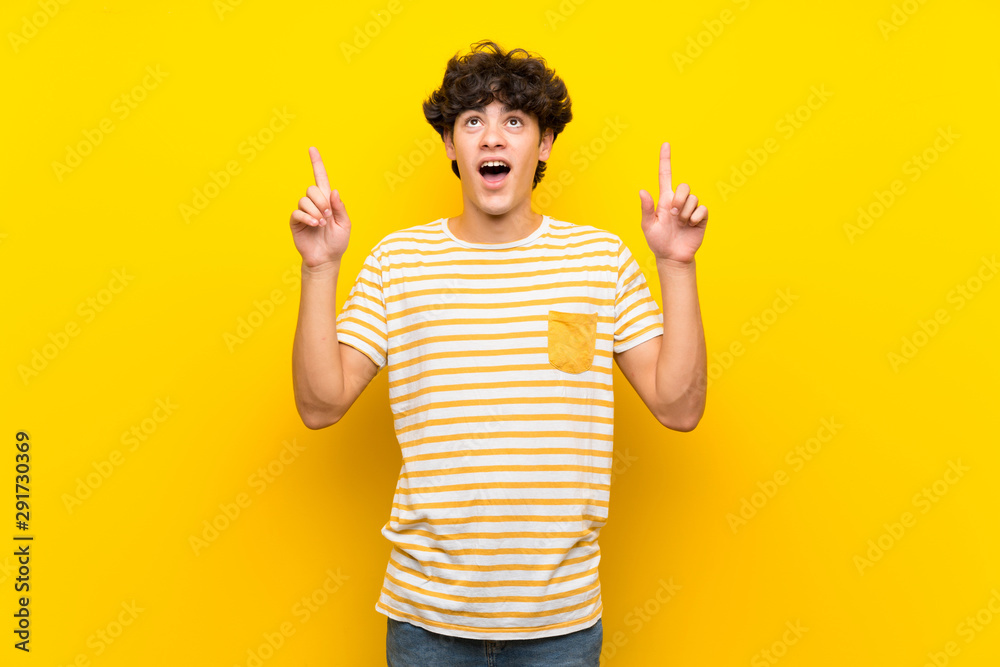 Young man over isolated yellow wall surprised and pointing up Young man over isolated yellow wall surprised and pointing up