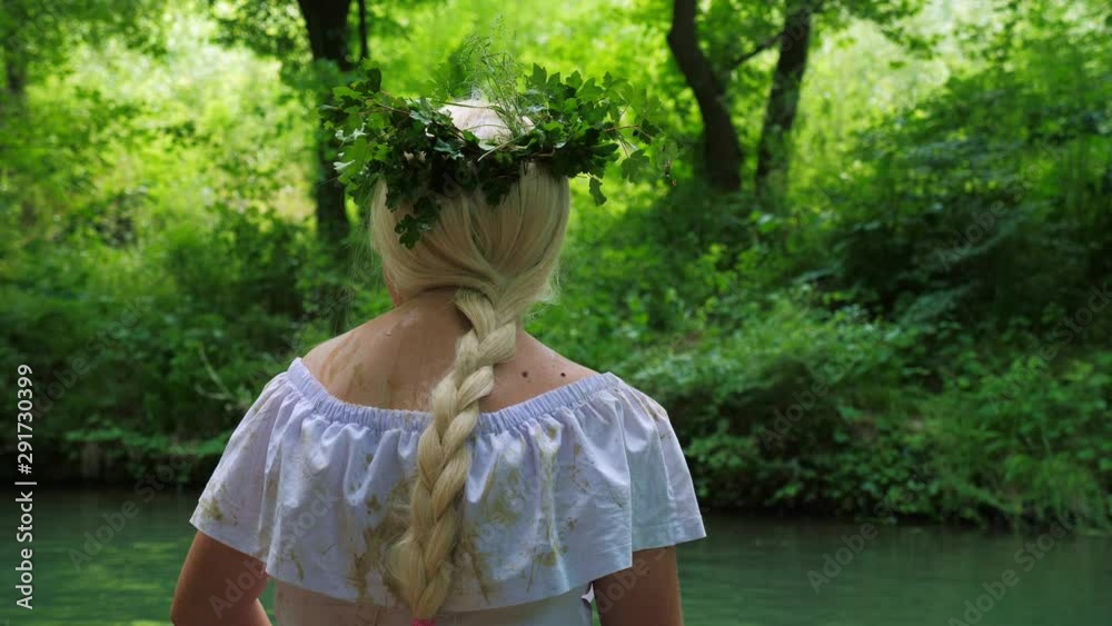 Unrecognizable girl blonde in white dirty clothes and a wreath of plants sitting on the banks of a forest river. Summer time. Unrecognizable girl blonde in white dirty clothes and a wreath of plants sitting on the banks of a forest river. Summer time.