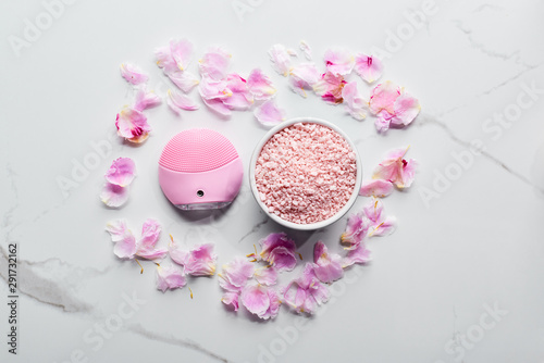 top view of silicone cleansing facial brush and bath salts on marble surface with pink petals