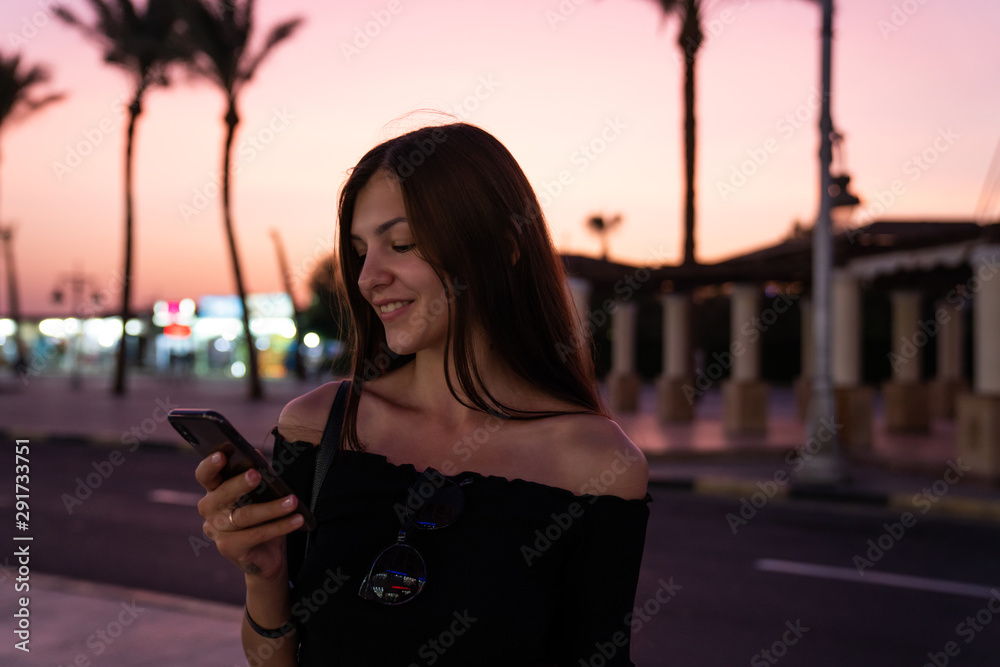 Fototapeta premium Happy brunette girl in black top reading message on her phone, blurred street with palms on the baclground