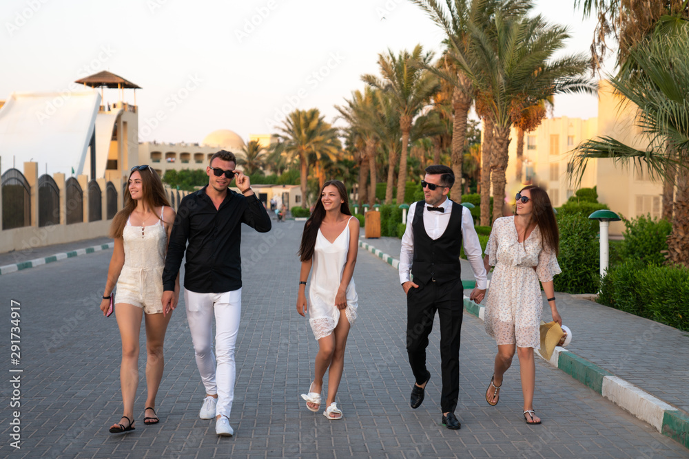 Fototapeta premium Five young friends in elagant clothes walking down the street with palms