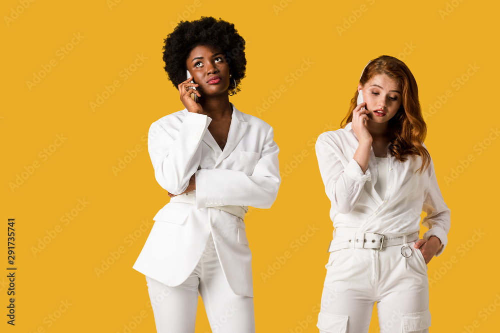  multicultural girls talking on smartphones while standing isolated on orange