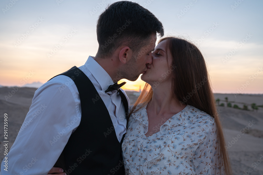 Beautiful young couple in elegant clothes kissing at sunset among the desert