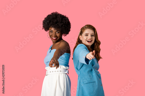 happy multicultural women pointing with fingers isolated on pink