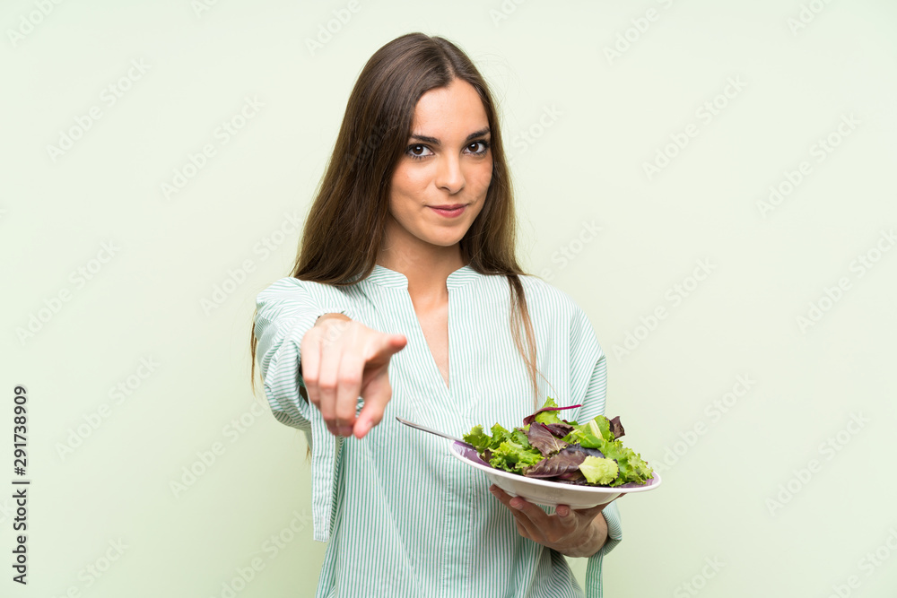 Young woman with salad over isolated green wall points finger at you with a confident expression Young woman with salad over isolated green wall points finger at you with a confident expression