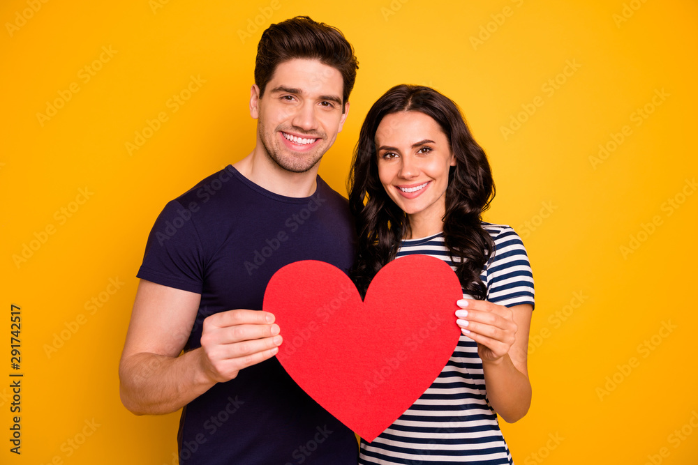 Photo of cheerful nice couple toothily smiling holding big red heart in love with each other while isolated with yellow background