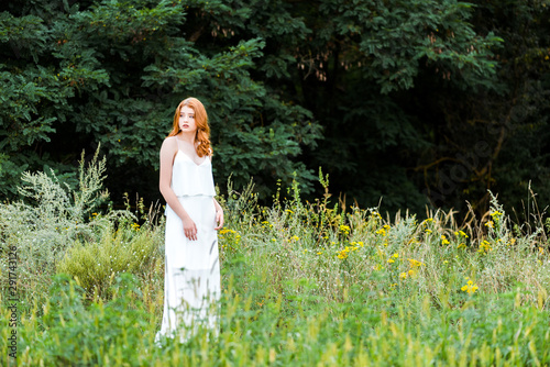 selective focus of beautiful redhead girl in white dress standing in field with wildflowers