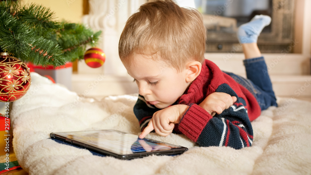 Naklejka premium Portrait of little boy lying on blanket next to Christmas tree and playing on digital tablet computer