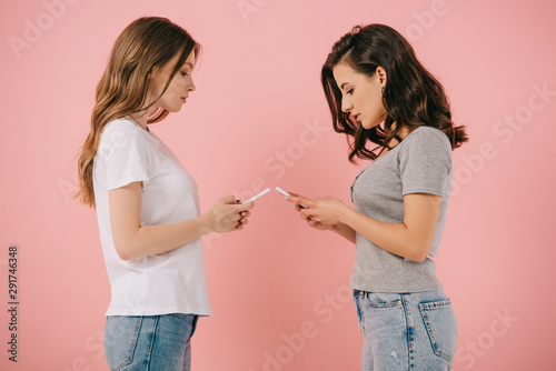 side view of attractive women in t-shirts using smartphones on pink background