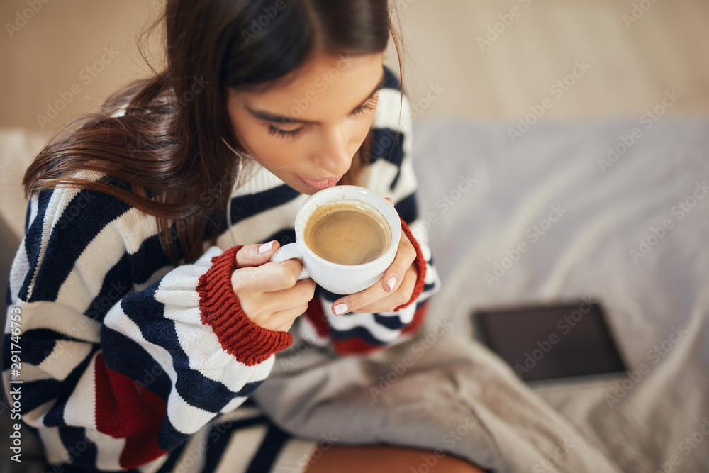 Top view of beautiful caucasian brunette in striped sweater sitting on bed in bedroom and drinking her morning coffee.