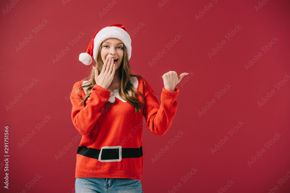 attractive woman in santa hat and sweater pointing with finger isolated on red