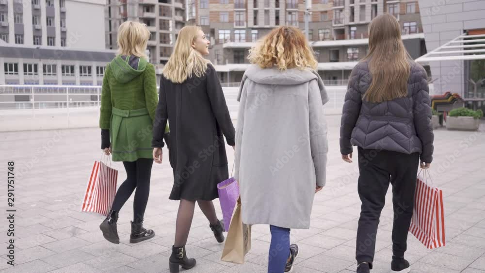 Back view beautiful girlfriends walking on the street talking happily. Four fashion women with shopping bags walking in the city. Leisure together. Girls enjoying their purchases Back view beautiful girlfriends walking on the street talking happily. Four fashion women with shopping bags walking in the city. Leisure together. Girls enjoying their purchases