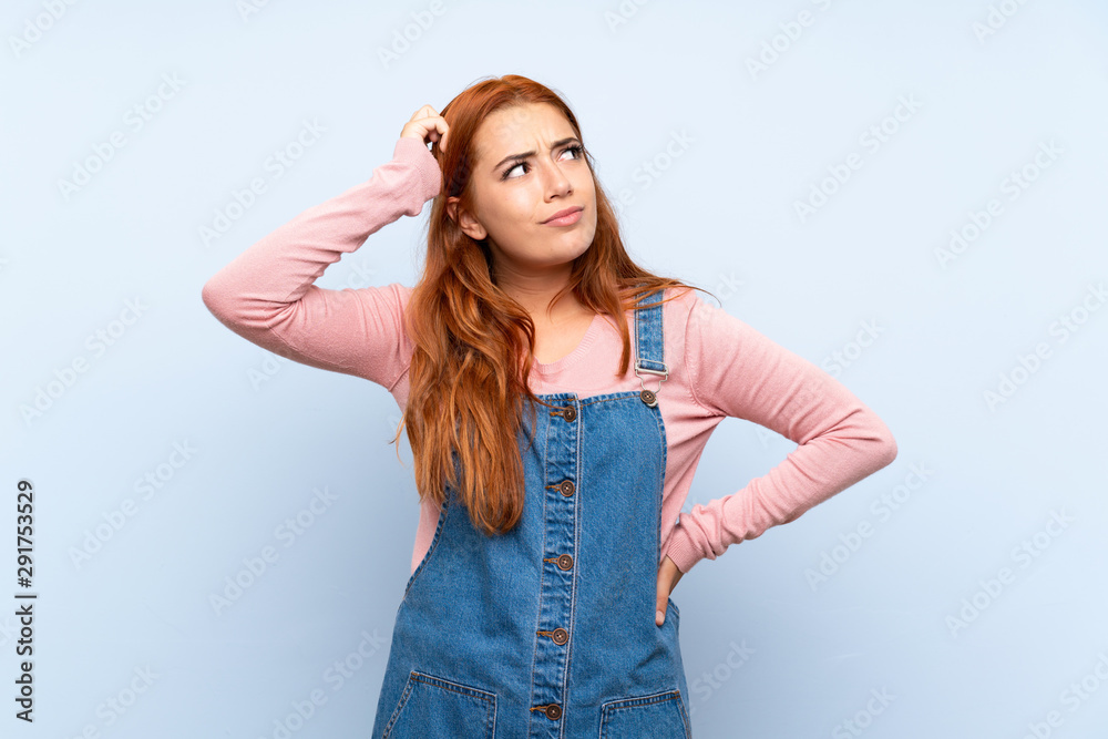 Teenager redhead girl with overalls over isolated blue background having doubts and with confuse face expression Teenager redhead girl with overalls over isolated blue background having doubts and with confuse face expression
