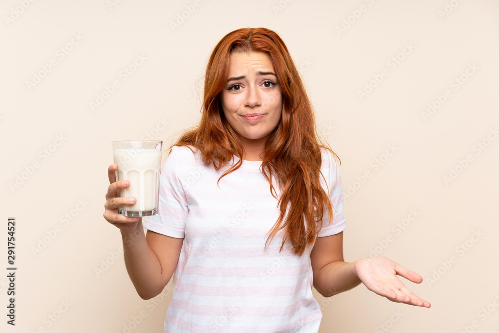 Teenager redhead girl holding a glass of milk over isolated background making doubts gesture while lifting the shoulders Teenager redhead girl holding a glass of milk over isolated background making doubts gesture while lifting the shoulders