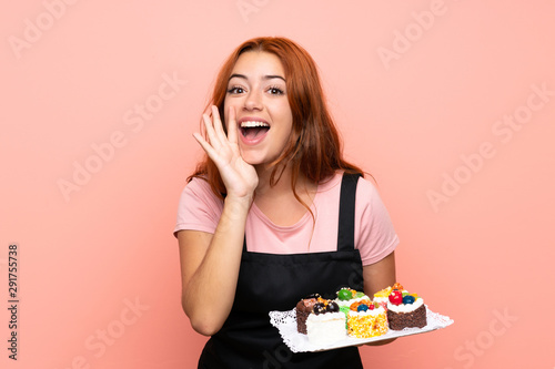 Teenager redhead girl holding lots of different mini cakes over isolated pink background shouting with mouth wide open