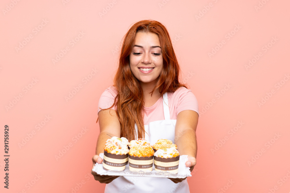 Teenager redhead girl holding lots of different mini cakes over isolated pink background Teenager redhead girl holding lots of different mini cakes over isolated pink background