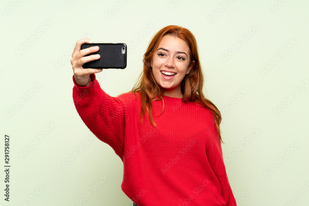 Teenager redhead girl with sweater over isolated green background making selfie with cellphone Teenager redhead girl with sweater over isolated green background making selfie with cellphone