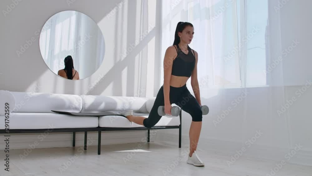 Brunette in black sportswear in a white apartment makes a split squat with dumbbells in her hands leaning on the sofa with her foot Brunette in black sportswear in a white apartment makes a split squat with dumbbells in her hands leaning on the sofa with her foot