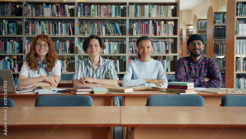 Multi-ethnic group of young people girls and guys students are sitting at desk in library smiling looking at camera. Lifestyle, education and youth concept. Multi-ethnic group of young people girls and guys students are sitting at desk in library smiling looking at camera. Lifestyle, education and youth concept.