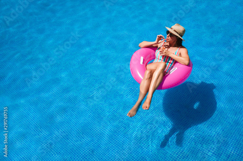 Young woman with sunglasses, hat and swimsuit in a blue pool. Pretty girl on a pink float enjoying the summer while having a cocktail.