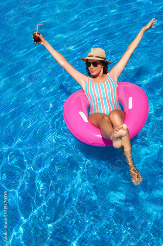 Young woman with sunglasses, hat and swimsuit in a blue pool. Pretty girl on a pink float enjoying the summer with open arms while having a cocktail.