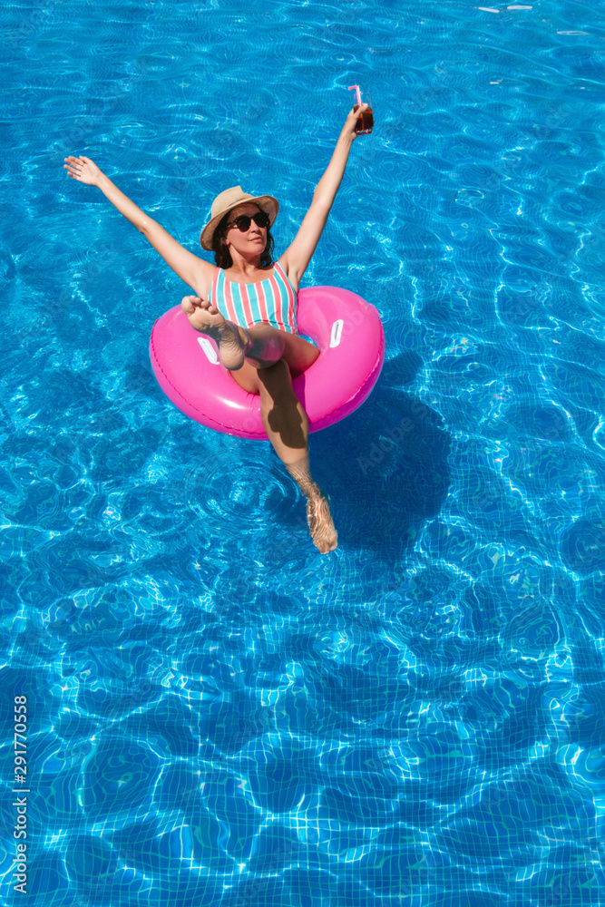 Young woman with sunglasses, hat and swimsuit in a blue pool. Pretty girl on a pink float enjoying the summer with open arms while having a cocktail.