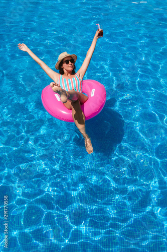 Young woman with sunglasses, hat and swimsuit in a blue pool. Pretty girl on a pink float enjoying the summer with open arms while having a cocktail.