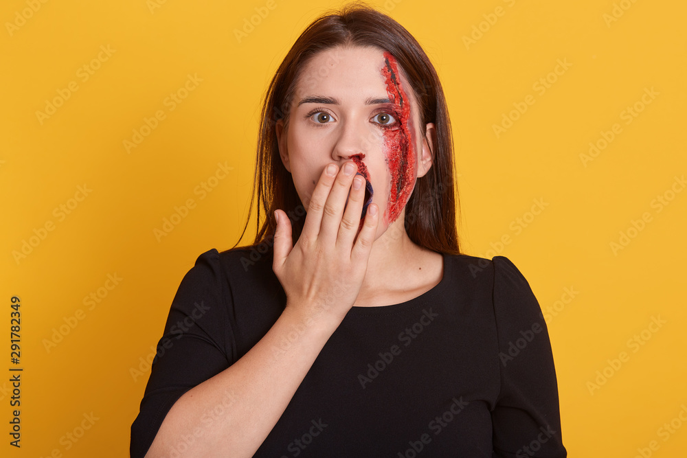 Indoor shot of beautiful young woman covering her mouth with hand, isolated over yellow background, has bloody wound on her cheek, looks shocked and astonished, dresses black shirt. Halloween concept. Indoor shot of beautiful young woman covering her mouth with hand, isolated over yellow background, has bloody wound on her cheek, looks shocked and astonished, dresses black shirt. Halloween concept.