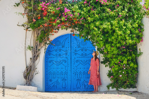 Fotografie, Obraz Woman next to home entwined liana with flowers in Sidi Bou Said, Tunisia - June