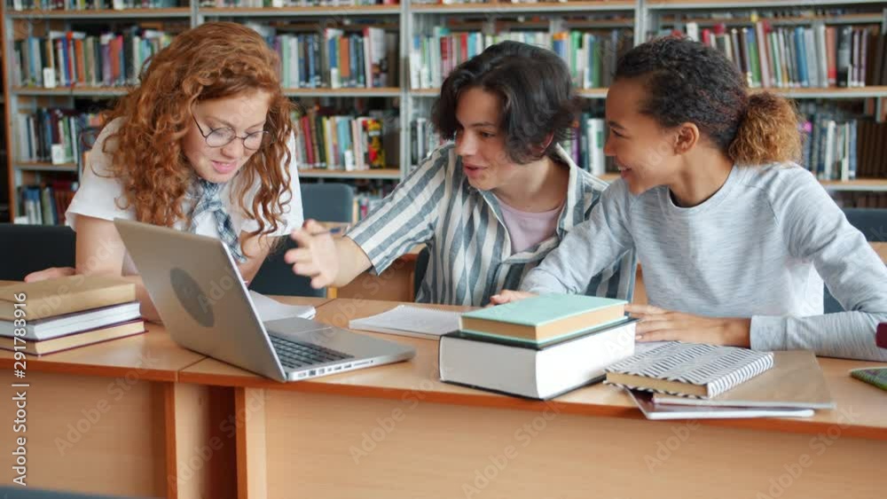 Cheerful students girls and guy working at project in library using laptop and books talking reading writing. Cooperation, teamwork and youth concept. Cheerful students girls and guy working at project in library using laptop and books talking reading writing. Cooperation, teamwork and youth concept.