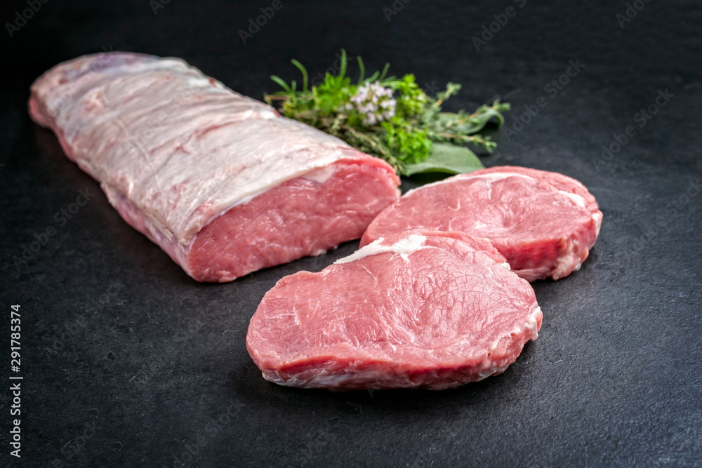 Roher dry aged Kalbsrücken angeboten mit Kräuter als closeup auf einem schwarzen Board mit Textfreiraum Roher dry aged Kalbsrücken angeboten mit Kräuter als closeup auf einem schwarzen Board mit Textfreiraum