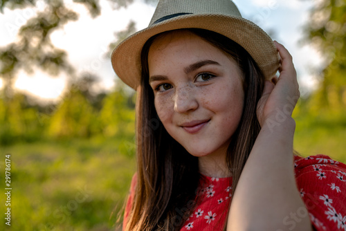 Close up portrait of the cute teen read head girl with freckles dressed in red dress among the garden touching her hat with her hand and looking at the camera