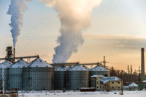 Elevator for grain storage and agricultural complex against the background of smoking chimneys. Evening winter photo.