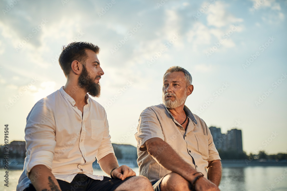 Father and adult son sitting on a wall at the riverside talking Father and adult son sitting on a wall at the riverside talking