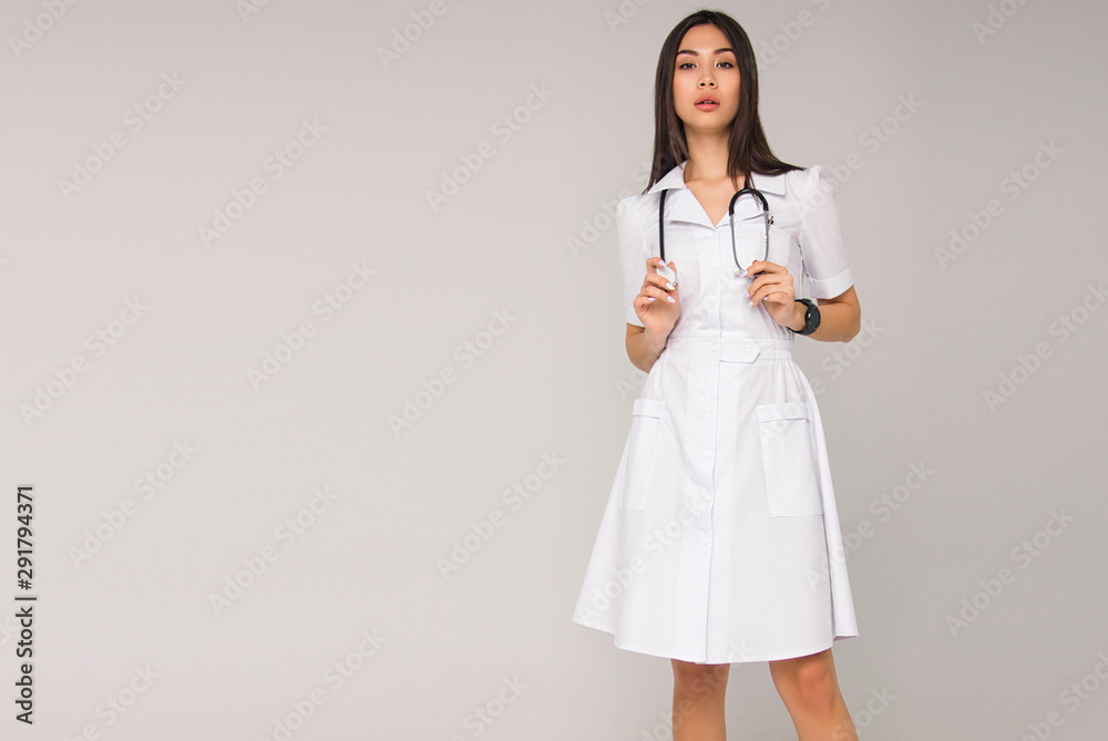 young stylish brunette nurse with long hair in white medical dress with stethoscope on her neck is standing on the white wall background. medical fashion concept. free space on left side young stylish brunette nurse with long hair in white medical dress with stethoscope on her neck is standing on the white wall background. medical fashion concept. free space on left side