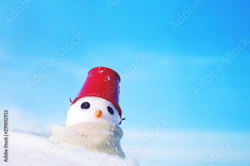 Little snowman made of snow in scarf and red bucket on his head on snow on blue sky background, soft focus. Christmas background