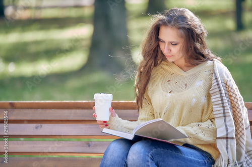 woman in jeans and a sweater sits in an autumn park with a thermocup in her hands and reads a book photo