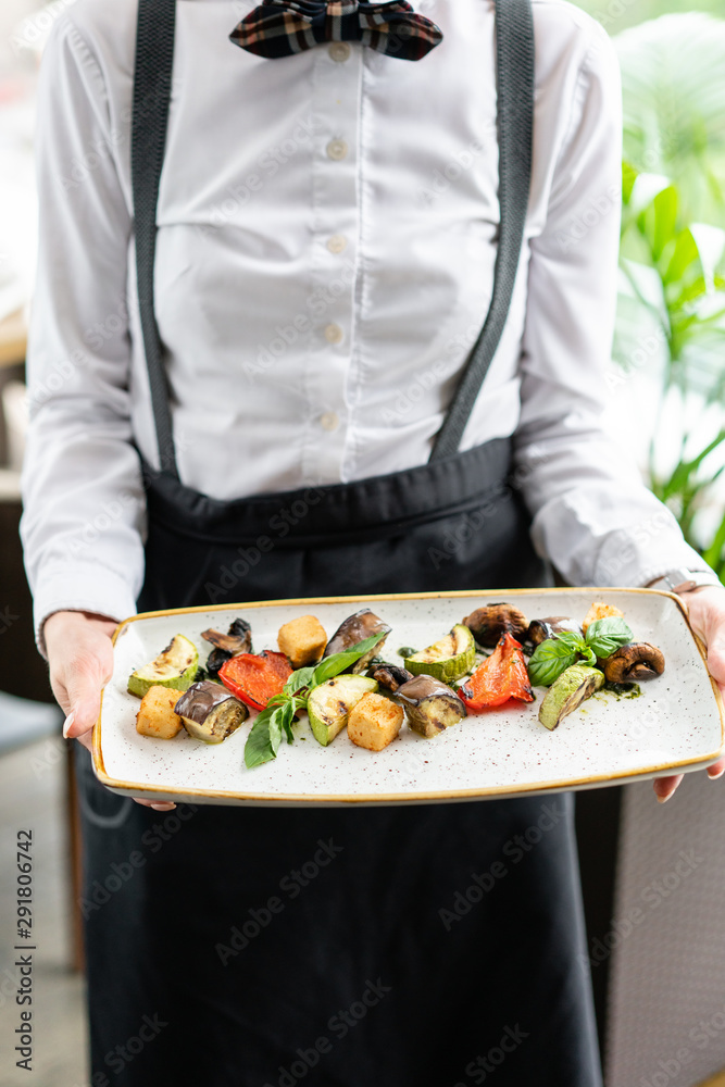 The waiter is holding a plate Grilled vegetables with feta cheese. Serving on a plate on a table. Barbecue restaurant menu, a series of photos of different dishes The waiter is holding a plate Grilled vegetables with feta cheese. Serving on a plate on a table. Barbecue restaurant menu, a series of photos of different dishes