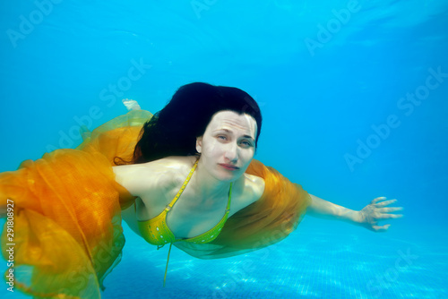 A grown girl swims underwater near the bottom of the pool with her long hair down and a yellow cloth over her shoulders on a Sunny day. She looks at the camera. Surreal portrait