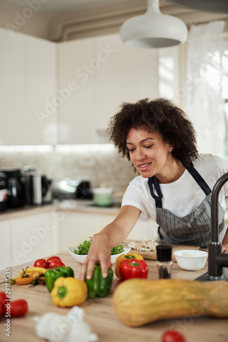 Beautiful mixed race housewife reaching for green pepper while standing in kitchen. Dinner preparation.