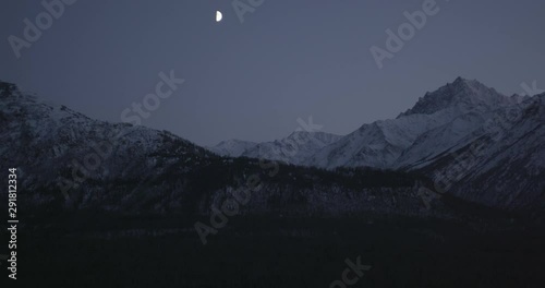  Aerial overhead helicopter shot, half moon gleams in between rocky and snowy Alaskan peaks, drone footage