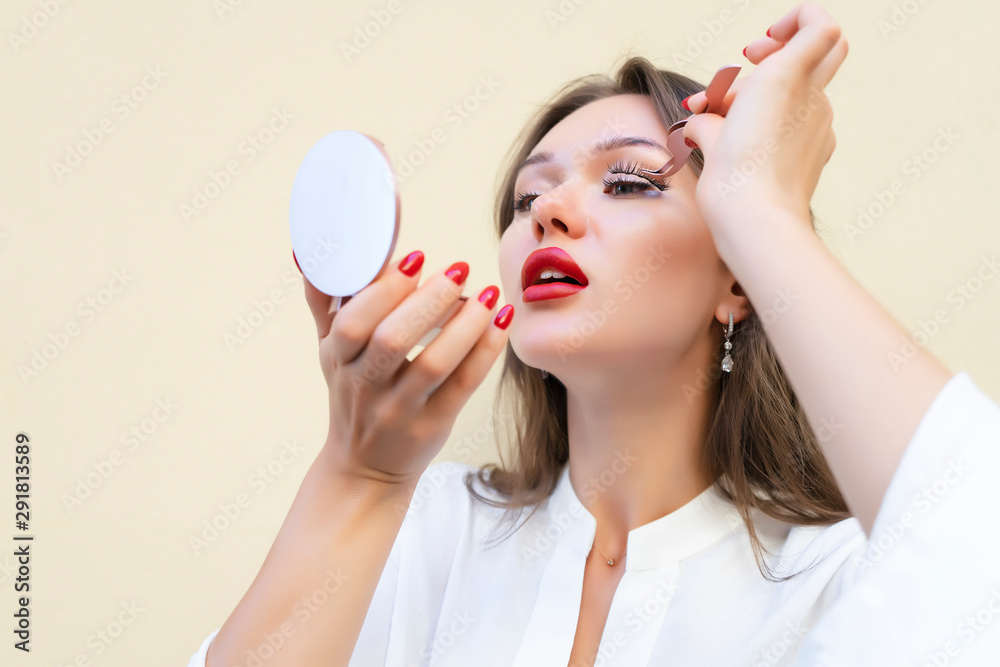 Charming woman with big red lips in white blouse applying magnetic fake eyelashes isolated on beige color. Beauty treatment concept for cosmetologist. Pure perfect skin. Selective focus. Copy space Charming woman with big red lips in white blouse applying magnetic fake eyelashes isolated on beige color. Beauty treatment concept for cosmetologist. Pure perfect skin. Selective focus. Copy space