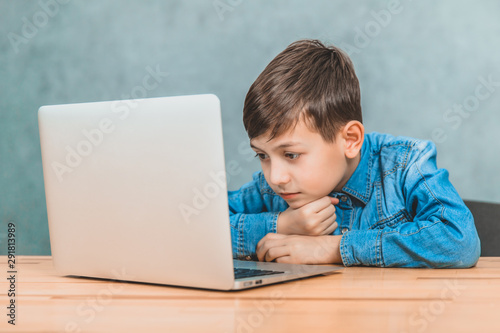 Cute little schoolboy sitting at the desk with the laptop, searching in the internet.